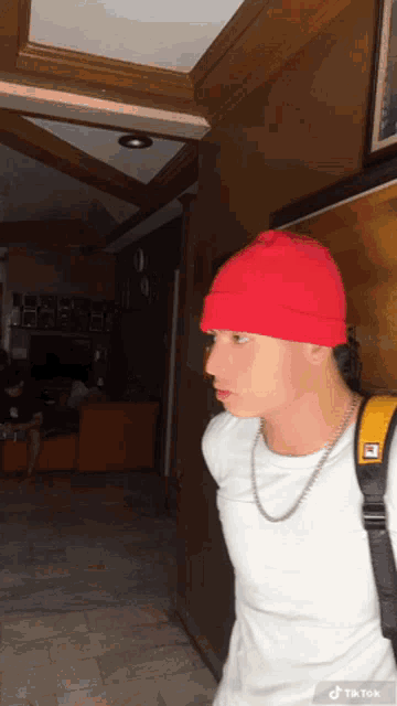 a young man wearing a red beanie and a white shirt is walking down a hallway with a backpack ..