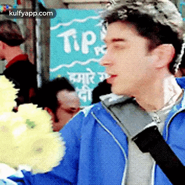 a man in a blue jacket is holding a bouquet of yellow flowers in front of a sign that says tip