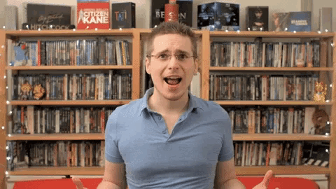 a man in a blue shirt stands in front of a shelf full of dvds including hitchcock and citizen kane