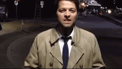 a man in a trench coat and tie is standing in front of a street at night .