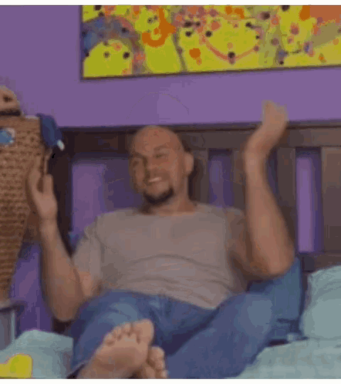 a bald man with a beard is sitting on a bed with his arms crossed .