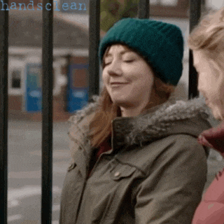 a woman wearing a green hat and a parka stands next to a fence with the words handsclean written on it