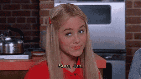 a blonde woman in a red shirt is sitting in a kitchen talking to someone .