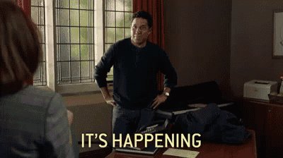a man is standing in front of a woman in a room with the words `` it 's happening '' written on the wall .