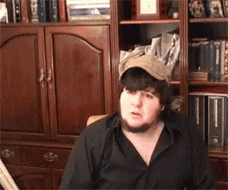 a man wearing a hat and a black shirt is sitting in front of a bookcase