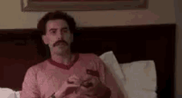 a man with a mustache is sitting on a bed holding a cell phone .
