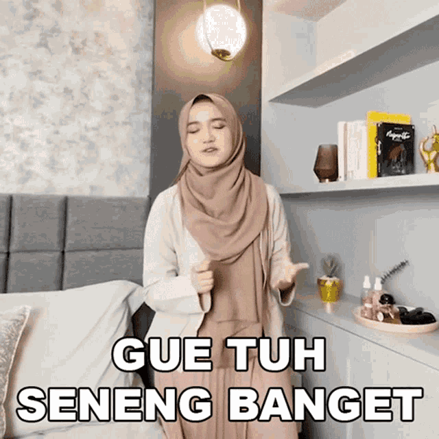 a woman in a hijab is standing in front of a bed with the words gue tuh seneng banget above her