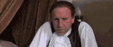 a man in a white shirt and black vest is sitting in a room with a curtain behind him .