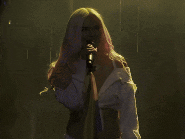 a woman with pink hair singing into a microphone with a yellow and blue flag behind her