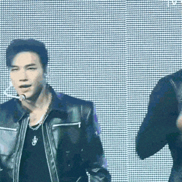 a man in a leather jacket is singing into a microphone while another man stands behind him .