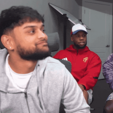 a man in a red jacket with the word manchester on it sits next to another man