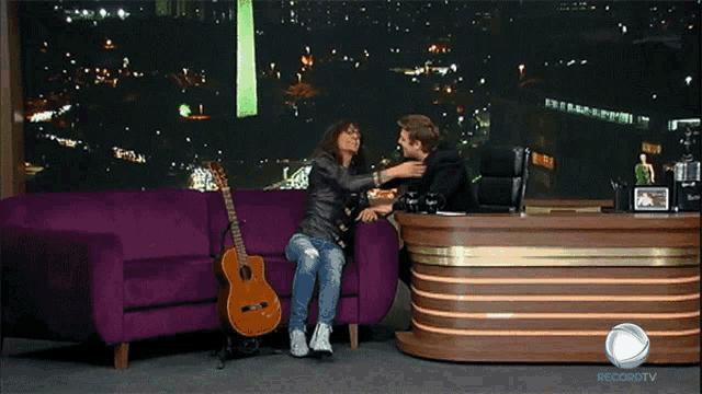 a man sitting on a purple couch talking to a woman with a guitar