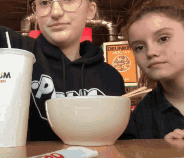 two girls are sitting at a table with a drink and a bowl of noodles