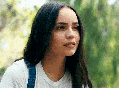 a woman with long dark hair is wearing a white shirt and denim overalls and looking at the camera .