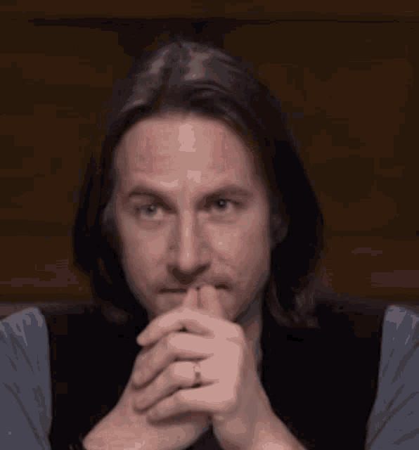 a man with long hair and a ring on his finger is sitting with his hands folded .