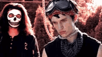 a man wearing a skull mask stands next to another man wearing a bandana and goggles