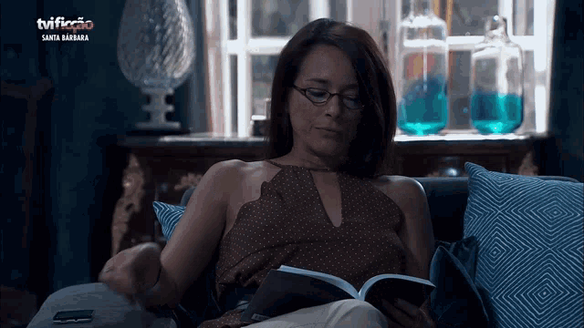 a woman is sitting on a couch reading a book and the tv shows santa barbara on the screen behind her