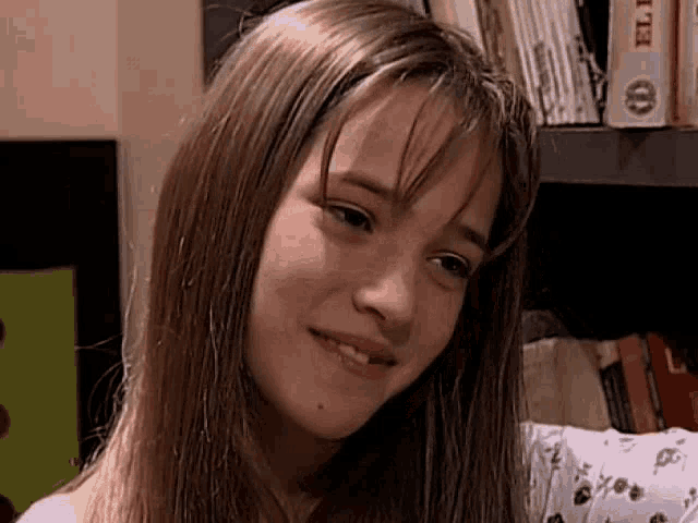 a young girl with long hair is smiling in front of a bookshelf with books on it .