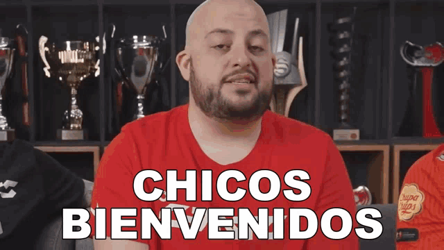 a man in a red shirt says chicos bienvenidos in front of trophies