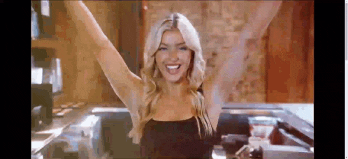 a woman in a black top is standing in a kitchen with her arms in the air .