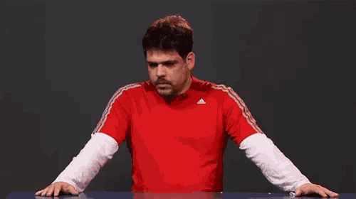 a man in a red adidas shirt is standing at a table with his hands on the table .