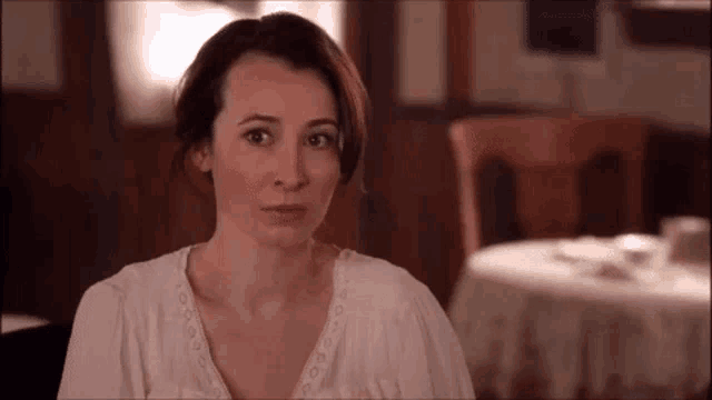 a woman in a white shirt is sitting at a table in a restaurant and looking at the camera .