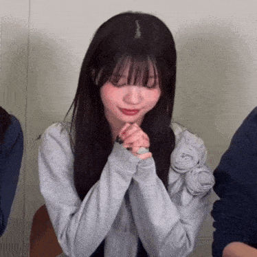 a girl with her hands folded in front of her face is wearing a grey sweater