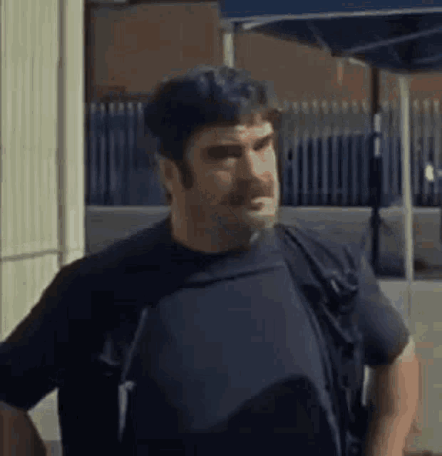 a man with a beard and mustache is standing in front of a building with his arms crossed .