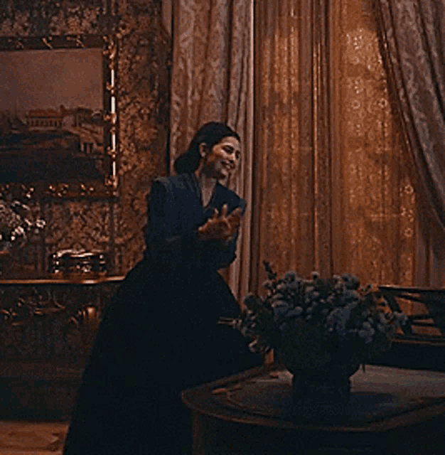 a woman in a black dress is standing in a room with a vase of flowers