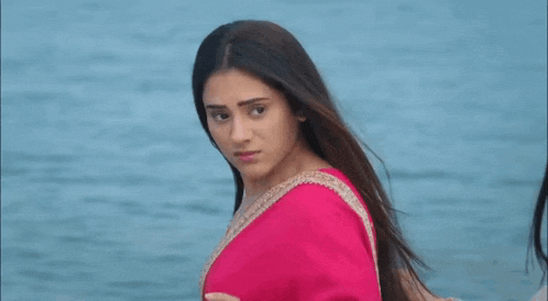 a woman in a pink saree is standing in front of the ocean looking at the camera .