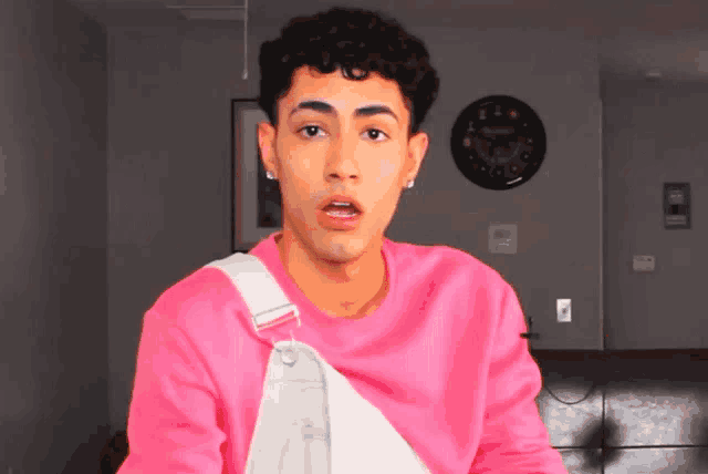 a young man wearing a pink shirt and white overalls is looking at the camera