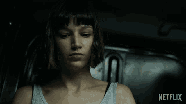 a woman in a grey tank top is sitting in a dark room with a netflix logo in the corner