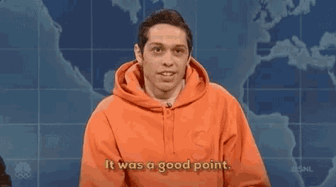 a man in an orange hoodie is standing in front of a map of the world .
