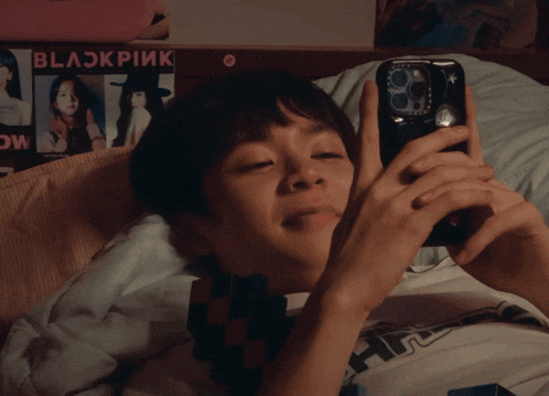 a person laying on a bed looking at a cell phone with a blackpink sticker on the wall behind them