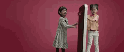 two little girls are standing next to each other on a pink background holding a large object .