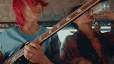 a woman with red hair is playing a guitar while another woman drinks a can of soda .