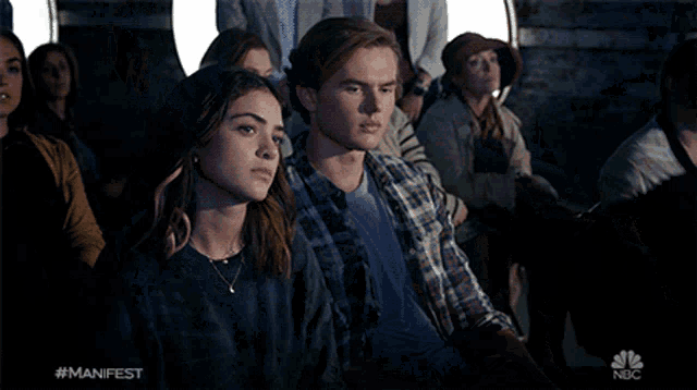a man and a woman sit in a crowd with #manifest on the bottom