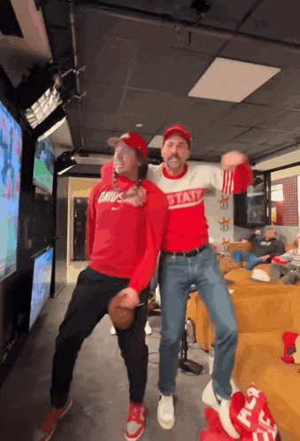 two men are dancing in a room with one wearing a sweater that says state