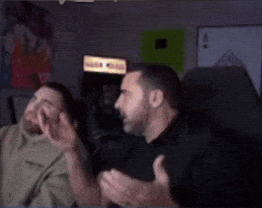 two men are sitting in front of an arcade machine and talking to each other .