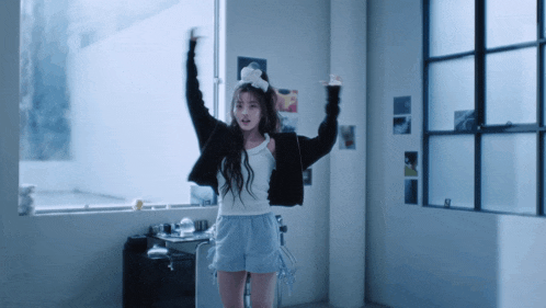 a woman in a white tank top and blue shorts stands in a room with her arms in the air