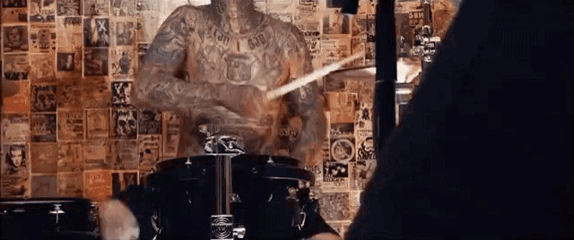 a man with a lot of tattoos is playing drums in front of a wall with posters on it .