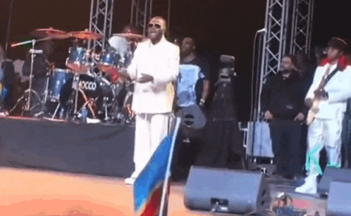 a man in a white suit stands in front of a drum set that says xcco