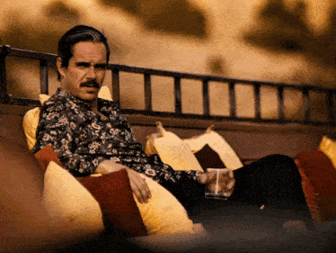 a man with a mustache sits on a couch with a glass of whiskey