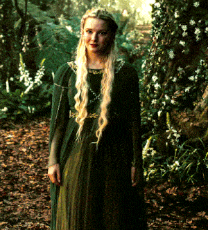 a woman in a green dress and cape stands in the woods