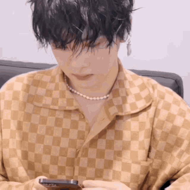 a man wearing a checkered shirt and a pearl necklace is looking at his cell phone