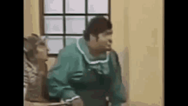 a man in a green shirt is standing in front of a window and talking on a cell phone .
