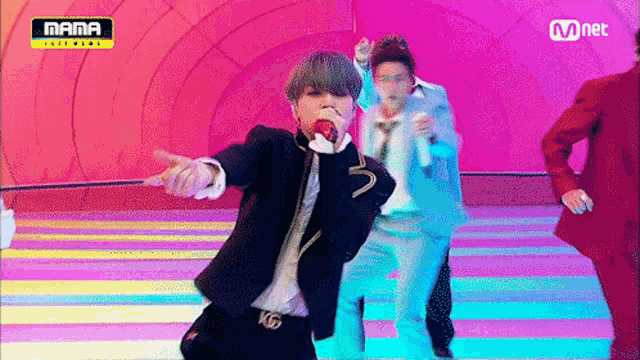 a man in a suit is singing into a microphone while two other men are dancing in front of a mnet banner