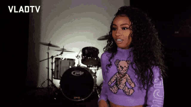 a woman is standing in front of a drum set wearing a purple sweater with a teddy bear on it .