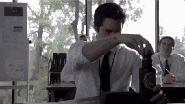 a man in a white shirt and tie is sitting at a desk in a classroom holding a water bottle .