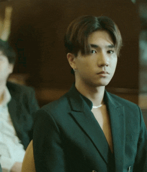 a young man wearing a green suit and a pearl necklace looks at the camera